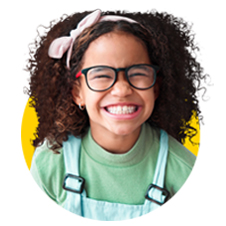 A older child with curly hair and a huge smile wearing overalls, thick framed glasses, and a pink headband with a bow.